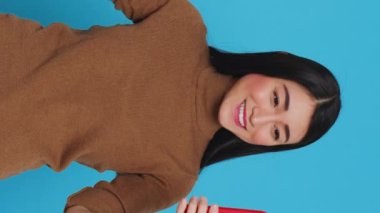 Vertical video: Smiling housekeeper ironing clothes using steamer while having videocall meeting on smartphone, talking with remote friend. Asian housewife committed to maintaining high standards of