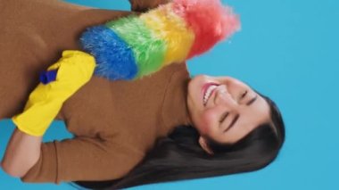 Vertical video: Smiling housekeeper holding colorful feather dust while taking selfie with mobile phone, standing in studio over blue background. Cheerful maid using high-quality detergents and rubber