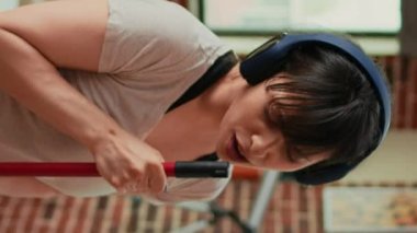 Vertical video: Cheerful girlfriend listening to music on headset and mopping floors, sweep dirt. Housewife feeling happy and cleaning household with tools and appliances, enjoying dance moves.