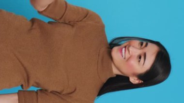Vertical video: Smiling cleaning lady using steamer to ironing clothes in studio over blue background. Woman used the best cleaning products and equipment to ensure a sparkling, fresh-smelling home