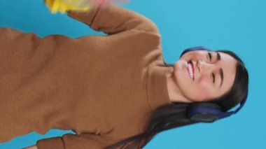 Vertical video: Cheeful housekeeper wearing headphones while dancing and having fun during house cleaning using feather duster. Overwhelmed maid role was vital to the smooth operation and cleanliness