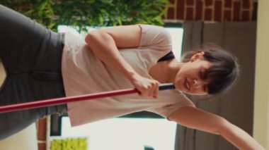 Vertical video: Young woman singing and dancing in living room, washing apartment floors with mop and washing solution. Cheerful housewife mopping tiles and having fun spring cleaning. Handheld shot.