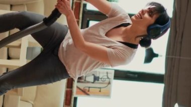 Vertical video: Happy housewife listening to music on headphones and vacuuming floors, having fun with vacuum cleaner. Young smiling person feeling positive while she does spring cleaning at home