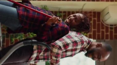 Vertical video: Happy partners showing silly dance moves and cleaning apartment, mopping and vacuuming floors while they listen to music. Modern positive people using mop and vacuum cleaner. Handheld