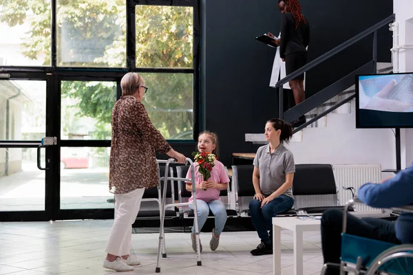 Granddaughter giving flowers to grandmother after finishing medical examination with practitioner doctor. Elderly patient with walking frame discussing disease expertise with family