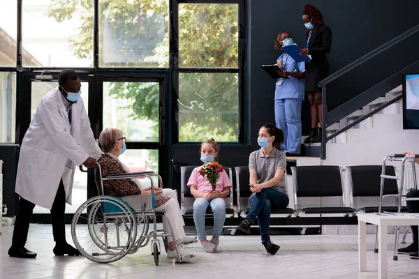 Physician doctor bringing grandmother in wheelchair to family after medical consultation during checkup visit appointment. Granddaughter holding bouqet flowers for senior woman. Virus epidemic