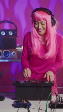Vertical video: Smiling artist standing at dj table enjoying mixing stereo sounds with electronics, having fun in club at night. Asian performer enjoying to perform music using professional audio