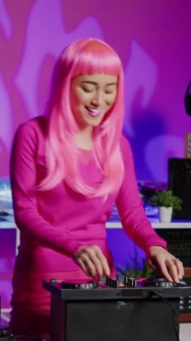 Vertical video: Smiling musician working as dj playing at mixer console, mixing techno sound with eletronic using audio equipment. Performer with pink hair having fun performing in club at night time