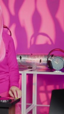 Vertical video: Artist having fun mixing eletronic music with techno using professional mixer console, standing at dj table in club at night. Musical performer with pink hair and blouse enjoying