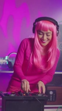Vertical video: Musician enjoying performing eletronic music at party in nightclub, using professional audio equipment. Asian performer with pink hair mixing techno sound using mixer console. Dj