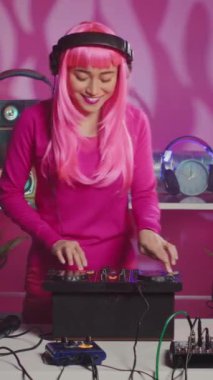 Vertical video: Musician with pink hair standing at dj table enjoying mixing stereo sounds with electronics, having fun in club at night. Smiling artist enjoying to perform music using professional