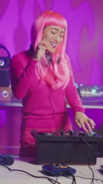 Vertical video: Smiling musician working as dj playing at turntables, mixing eletronic sound with techno using professional audio equipment. Asian artist with pink hair having fun performing in club