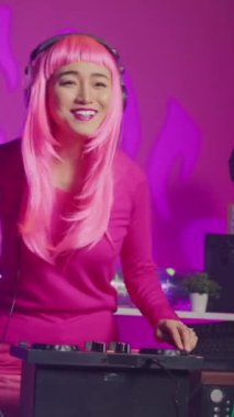 Vertical video: Musician standing at dj table performing electronic song using professional turntables, enjoying playing music during night time in club. Artist with pink hair doing performance with