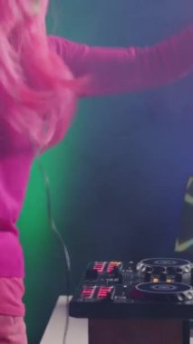 Vertical video: Smiling artist standing at dj table playing electronic music at professional mixer console in studio over pink background. Asian musician performing techno sound, having fun in club at