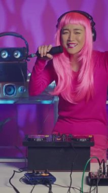 Vertical video: Musician talking with fans into microphone during party, performing techno remix using professional turntables. Asian performer mixing sounds while dancing and having fun in nightclub