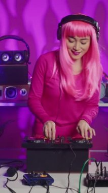 Vertical video: Cheeful artist standing at dj table playing electronic music using professional mixer console, enjoying to perform at night in club. Asian musician having fun with fans during techno