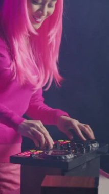 Vertical video: Smiling artist working as dj playing at mixer console, mixing techno sound with eletronic using professional audio equipment. Musician with pink hair having fun while performing in