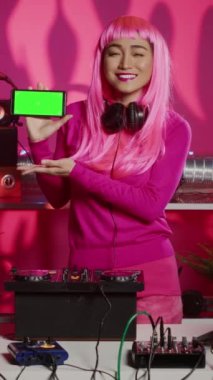 Vertical video: Dj artist presenting green screen mock up mobile phone with isolated display during musical performance in night club. Musician playing stereo sounds with electronics using