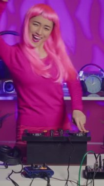 Vertical video: Asian performer dancing and interacting with fans in club at nightime, playing electronic remix at professional mixer console. Artist with pink hair mixing sounds using audio equipment
