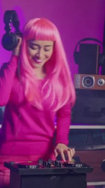 Vertical video: Musician with pink hair playing eletronic song at mixer console, listening music into headset. Asian dj perfomer enjoying mixing sounds, dancing with fans during night time in club.