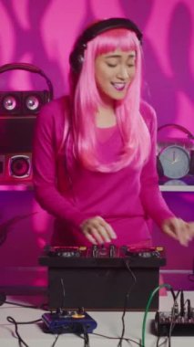 Vertical video: Asian performer playing electronic music at mixer console, having fun with fans in club during night time. Dj musician with pink hair mixing techno sound using professional audio