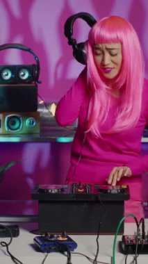 Vertical video: Cheerful musician playing techno music at turntables, enjoying having fun and dancing in club during party. Artist with pink hair performing remix sound using professional audio