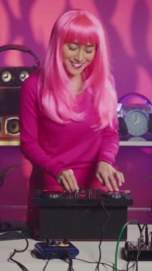 Vertical video: Artist with pink hair dancing and having fun mixing sound during night time in club, performing eletronic music using professional mixer console. Performer recording album using audio