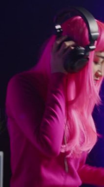 Vertical video: Dj with pink hair playing eletronic song at professional mixer console, listening music into headset. Asian perfomer enjoying mixing sounds, dancing with fans during night time in club