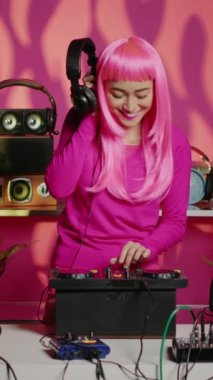 Vertical video: Smiling artist playing techno music at professional mixer while listening sound at headphones, having fun during concert in nightclub. Asian dj performing remix celebrating electronic