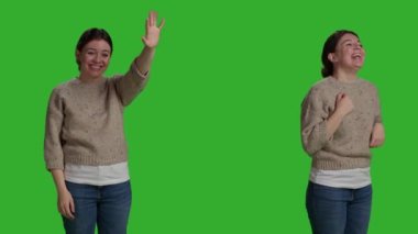 Close up of female model greeting someone and waving, smiling in studio. Relaxed young woman waving hi or hello on camera, standing over full body greenscreen backdrop and saying goodbye.