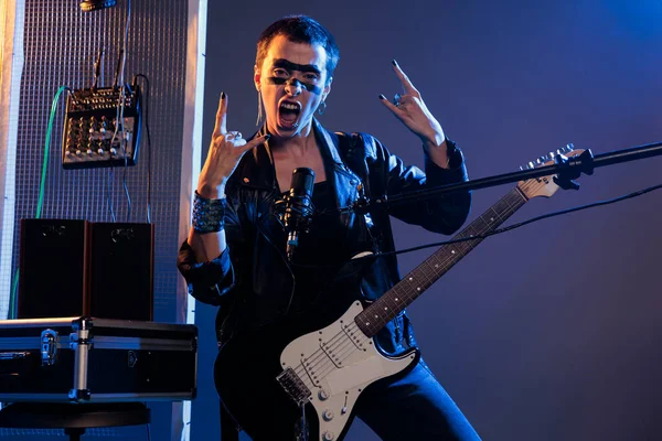 Punk rocker doing rock sign on live performance, singing loud alternative heavy metal music. Using microphone and guitar to play cool solo song, wearing leather jacket and showing rock on symbol.
