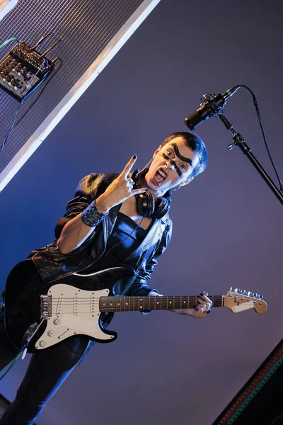 Punk rocker doing rock sign and playing guitar, feeling ecstatic and doing solo concert in studio over background. Female singer showing rock and roll symbol at live show performance.