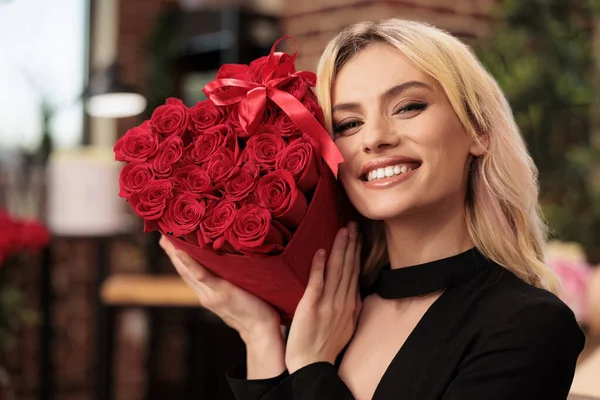 愛の休日を祝う赤いバラの花束を持っているガールフレンドは バレンタインデープレゼントでいっぱいのリビングルームに立っています 魅力的なかわいい女性でエレガントな黒のドレスを楽しんでロマンチックな贈り物 — ストック写真