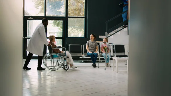 Old adult in wheelchair talking to young child in waiting area, family coming to visit senior patient with impairment. Wheelchair user trying to walk with walking frame in hospital lobby.