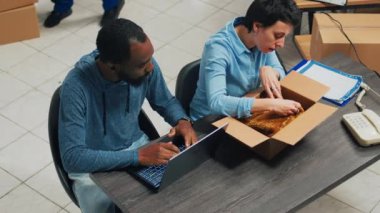 Man and woman preparing shipment order with merchandise from warehouse shelves, working on retail logistics with laptop. Diverse workers planning small business development, supply chain.