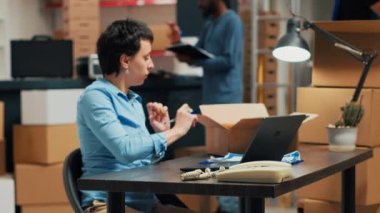 Female owner doing quality control before shipping goods in cardboard boxes, taking merchandise from warehouse shelves. Woman checking stock and doing distribution work. Handheld shot.