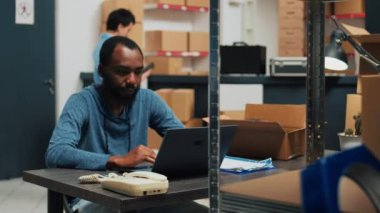 Male entrepreneur planning merchandise inventory and quality control, working on laptop to do retail logistics. Young man using stock in packages, shipping products and doing financial planning.