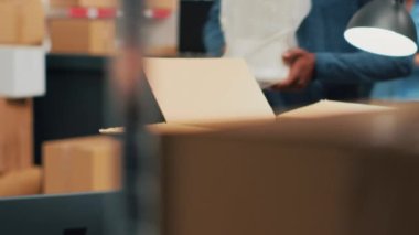 African american worker planning products shipment at warehouse, putting merchandise in boxes for distribution. Employee taking stock goods from storage shelves to ship supplies. Handheld shot.