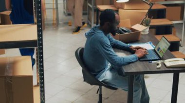 Young office worker checking goods in cardboard packages, looking at inventory stock on laptop in storehouse space. Business owner using merchandise from warehouse racks and shelves.