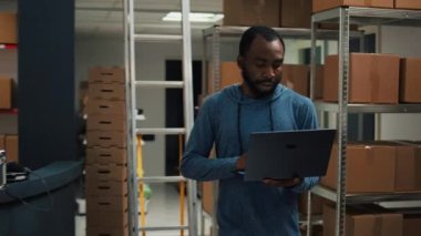 Young entrepreneurs doing teamwork to ship products in cardboard packages, checking quality of merchandise in storage room. Diverse people using laptop to plan goods shipment distribution.