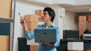 Small business owner using laptop to do storehouse inventory, planning quality control for products before shipping them. Person taking notes on pc and looking at merchandise packaging on shelves.