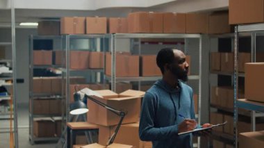 Male entrepreneur taking inventory notes on clipboard files, checking quality of goods or products and packages. Young employee looking at cardboard boxes, merchandise stock logistics.