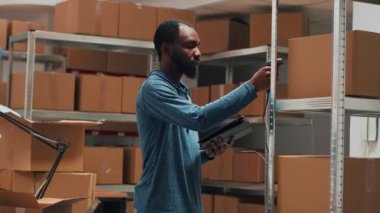 Male entrepreneur using scanner and tablet to check supplies inventory, working on products management. Young adult scanning merchandise in cardboard boxes on storehouse shelves. Handheld shot.