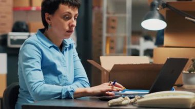 Young entrepreneur looking at merchandise in boxes to plan order shipment at storehouse space. Female adult using stock to do quality control and work on distribution management. Handheld shot.