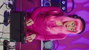 Vertical video: Musician with pink hair performing techno music using dj mixer console enjoying to play song with fans, having fun during night party. Artist doing performance with professional audio