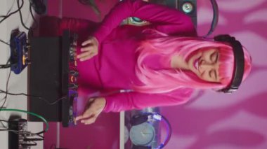 Vertical video: Musician with pink hair standing at dj table enjoying mixing stereo sounds with electronics, having fun in club at night. Smiling artist enjoying to perform music using professional