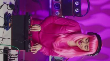 Vertical video: Cheeful artist standing at dj table playing electronic music using professional mixer console, enjoying to perform at night in club. Asian musician having fun with fans during techno