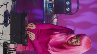 Vertical video: Asian musician playing electronic music at turntables, having fun with fans in club during night time. Dj artist with pink hair mixing techno sound using professional audio equipment
