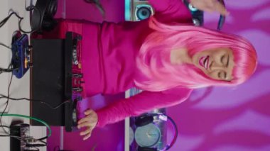 Vertical video: Cheerful performer dancing and interacting with fans in club at nightime, playing techno remix at professional mixer console. Artist with pink hair mixing electronic sounds using audio