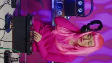 Vertical video: Asian musician playing electronic music at mixer console while having fun dancing in club during night time. Artist with pink hair performing remix sound using professional audio